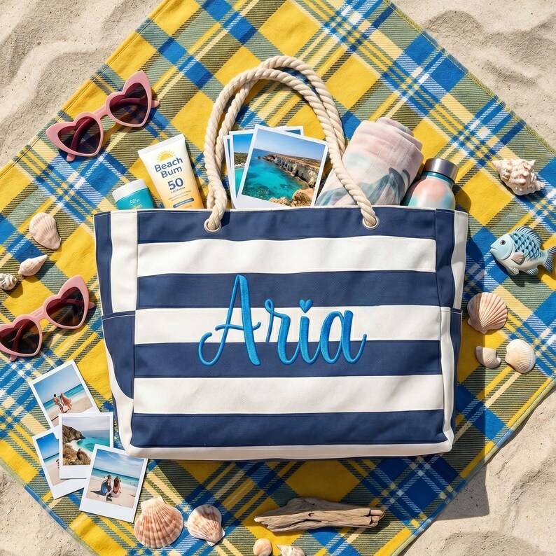 May include: A navy blue and white striped canvas tote bag with rope handles, featuring the name Aria embroidered in blue script on the front. The bag is filled with beach items including sunscreen, photos, and a towel, resting on a plaid blanket.