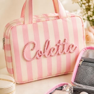 May include: A rectangular travel bag featuring vertical light pink and white stripes with two top handles. The name Colette is embroidered in a raised, textured pink rope script across the front. A silver zipper closure runs along the top edge.