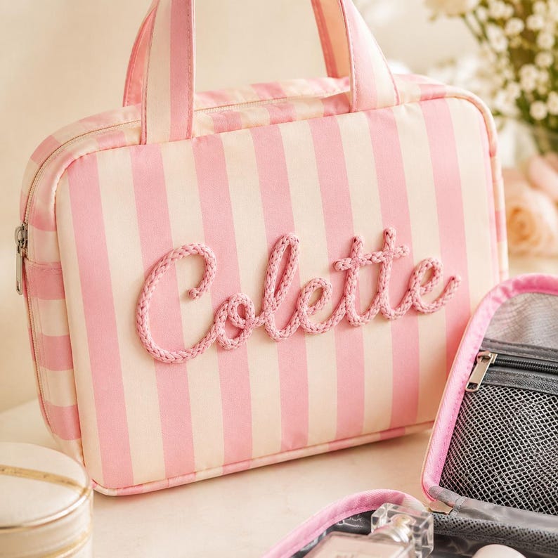 May include: A rectangular travel bag featuring vertical light pink and white stripes with two top handles. The name Colette is embroidered in a raised, textured pink rope script across the front. A silver zipper closure runs along the top edge.