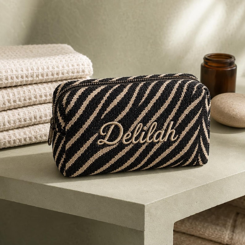 K&ouml;nnte beinhalten: Eine rechteckige Rei&szlig;verschlusstasche mit einem schwarz-beigen Zebramuster. Der Name Delilah ist in beiger Schreibschrift mittig auf der Vorderseite eingestickt. Die Tasche hat eine strukturierte Form mit einem Rei&szlig;verschluss oben.