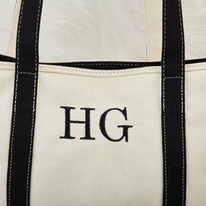 May include: Cream canvas tote bag with black handles and trim. The bag has the embroidered initials "HG" in black. The handles and trim are black, with white stitching. A practical and stylish bag for everyday use.
