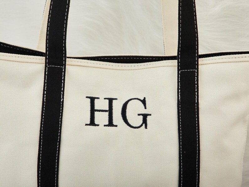May include: Cream canvas tote bag with black handles and trim. The bag has the embroidered initials "HG" in black. The handles and trim are black, with white stitching. A practical and stylish bag for everyday use.