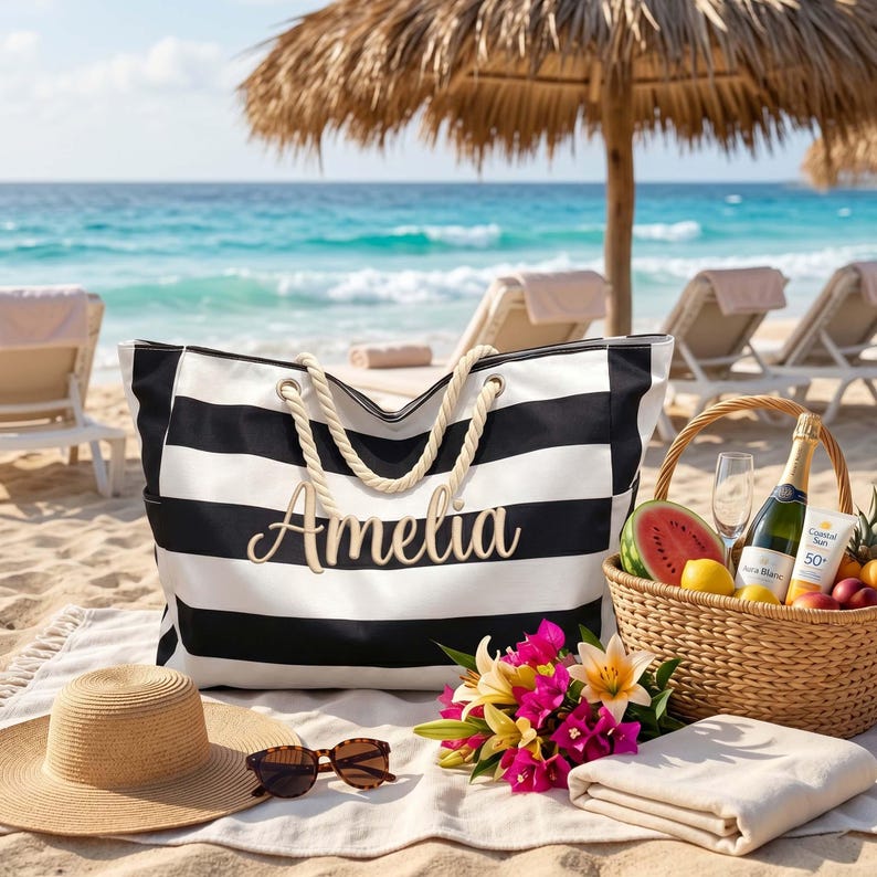 May include: A black and white striped canvas tote bag with thick rope handles and the name Amelia written in cursive script. The bag is placed on a white blanket alongside a straw hat, sunglasses, a wicker basket, and colorful flowers.