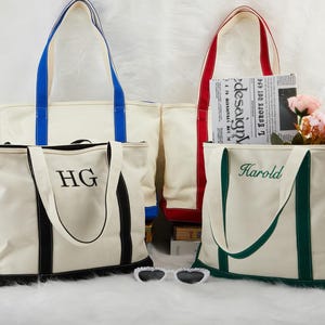 May include: Four cream canvas tote bags with coloured handles and trim. One bag has blue handles, another red, and a third green. The fourth bag has black trim and the initials "HG". One bag has the name "Harold" embroidered on it.
