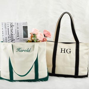 May include: Two canvas tote bags. One is white with green trim and the name "Harold" embroidered in green. The other is white with black trim and the initials "HG" embroidered in black. Both bags have long handles.