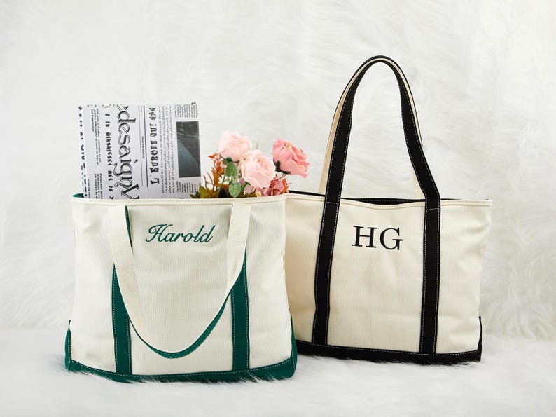May include: Two canvas tote bags. One is white with green trim and the name "Harold" embroidered in green. The other is white with black trim and the initials "HG" embroidered in black. Both bags have long handles.