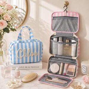 May include: A blue and white striped toiletry bag with the name Chloe embroidered on the front, alongside a matching pink and white striped hanging travel organizer filled with various skincare bottles, makeup compacts, and cosmetic brushes.