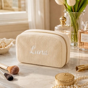 May include: A rectangular cream-colored corduroy cosmetic bag featuring a gold-tone zipper and the name Luna embroidered in white cursive script on the front. The bag has a textured ribbed surface and a structured shape for storing personal items.