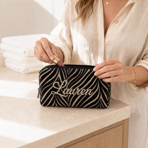 K&ouml;nnte beinhalten: Ein rechteckiges Rei&szlig;verschlussetui mit einem schwarz-beigen, zebraartigen Streifenmuster. Auf der Vorderseite ist der Name Lauren in einer kursiven Schriftart eingestickt. Das Etui hat eine strukturierte Form und einen schwarzen Rei&szlig;verschluss.