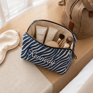 K&ouml;nnte beinhalten: Eine Kosmetiktasche mit Rei&szlig;verschluss und einem blau-beigen, zebraartigen Streifenmuster, auf deren Vorderseite der Name Kennedy in wei&szlig;er Schreibschrift eingestickt ist. Die offene Tasche enth&auml;lt zwei wei&szlig;e Hautpflege-Tuben und drei Pinsel.