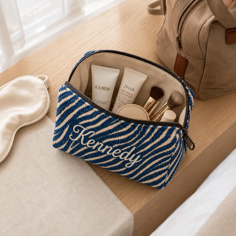 K&ouml;nnte beinhalten: Eine Kosmetiktasche mit Rei&szlig;verschluss und einem blau-beigen, zebraartigen Streifenmuster, auf deren Vorderseite der Name Kennedy in wei&szlig;er Schreibschrift eingestickt ist. Die offene Tasche enth&auml;lt zwei wei&szlig;e Hautpflege-Tuben und drei Pinsel.