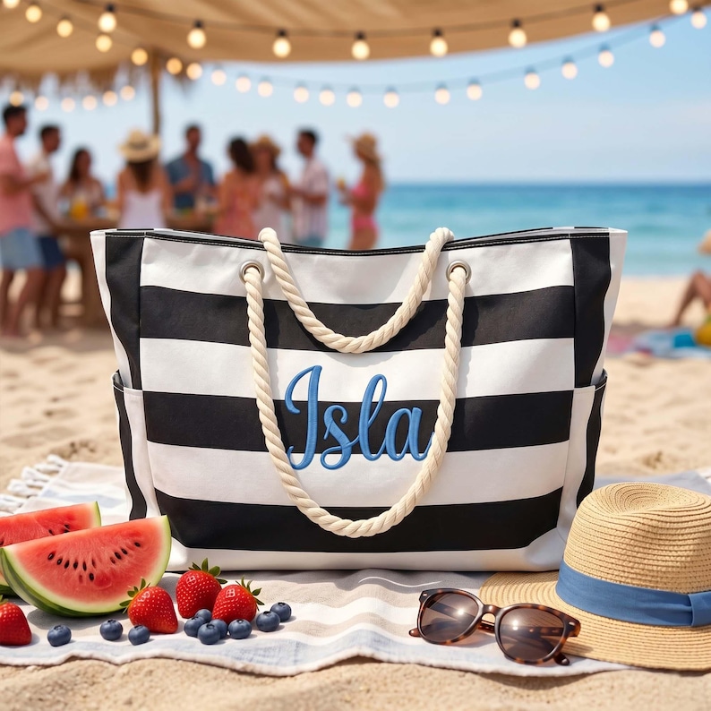 May include: A black and white striped tote bag with thick cream-colored rope handles. The front of the bag features the name Isla embroidered in blue script. The bag has side pockets and is constructed from a durable fabric material.
