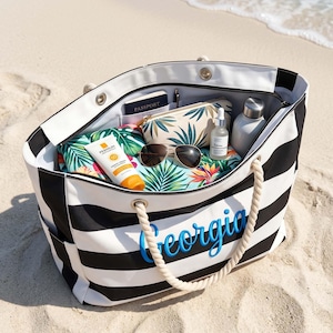 May include: A black and white striped tote bag with rope handles and the name Georgia embroidered in blue. The bag is filled with a passport, a tropical print pouch, sunglasses, a bottle of sunscreen, a dropper bottle, and a metal water bottle.