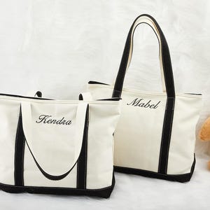 May include: Two cream canvas tote bags with black trim. The smaller bag has "Kendra" embroidered on it, and the larger bag has "Mabel". Both have black handles. Ideal for shopping or carrying essentials.