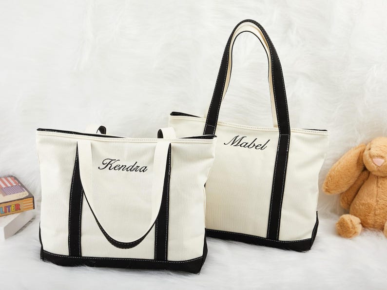 May include: Two cream canvas tote bags with black trim. The smaller bag has "Kendra" embroidered on it, and the larger bag has "Mabel". Both have black handles. Ideal for shopping or carrying essentials.