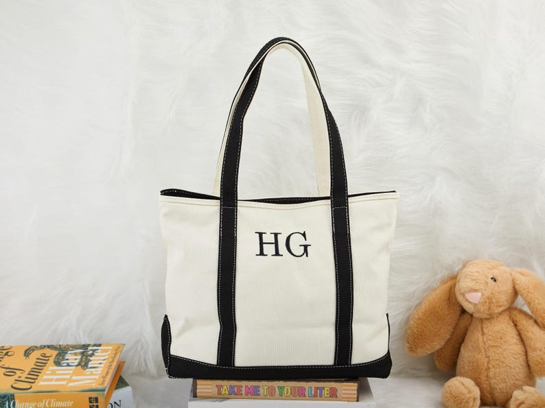 May include: Cream-coloured tote bag with black accents and handles. The bag features the embroidered initials "HG" in black. The bottom and trim are black. The bag is sitting on a wooden block with text.