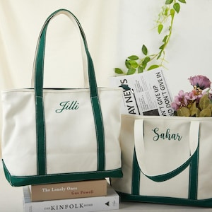 Könnte beinhalten: Zwei weiße Canvas-Tragetaschen mit grünen Akzenten. Auf einer Tasche ist "Jilli" gestickt, auf der anderen "Sahar". Die Taschen haben grüne Griffe und Böden. Eine Tasche enthält Blumen und eine Zeitung.