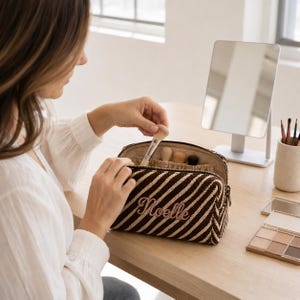 K&ouml;nnte beinhalten: Eine rechteckige Kosmetiktasche mit braun-cremefarbenem Diagonalstreifenmuster und dem in Schreibschrift gestickten Namen Noelle auf der Vorderseite. Die Tasche verf&uuml;gt &uuml;ber einen Rei&szlig;verschluss und Innenf&auml;cher f&uuml;r mehrere Make-up-Pinsel.