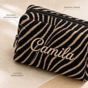 K&ouml;nnte beinhalten: Eine schwarz-beige Kosmetiktasche mit Zebramuster aus strukturiertem Webstoff, einem leichtg&auml;ngigen Rei&szlig;verschluss und dem Namen Camila in individueller Stickerei auf der Vorderseite. Das rechteckige Etui hat ein strukturiertes Design.