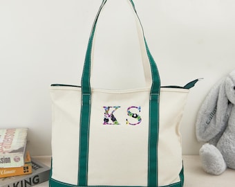 May include: A white canvas tote bag with green trim and a zipper closure. The bag has a floral embroidered design of the letters "KS" on the front.