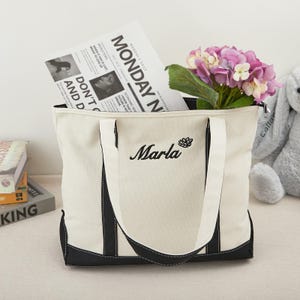 Könnte beinhalten: Cremefarbene Tragetasche mit schwarzen Akzenten und Griffen. Die Tasche trägt den Namen "Marla" in schwarzer Schrift gestickt, zusammen mit einem floralen Design. Im Inneren der Tasche befindet sich eine Zeitung mit der Überschrift "MONDAY N" und ein Strauß lila und weißer Blumen.