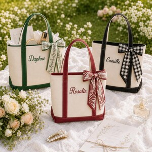 Pode incluir: Três sacolas de lona com alças superiores e laços decorativos. A bolsa da esquerda é verde com o nome Daphne, a bolsa central é bordô com o nome Rosalie e a bolsa da direita é preta com o nome Camille. Cada bolsa possui uma base sólida.