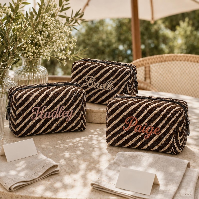 K&ouml;nnte beinhalten: Drei rechteckige Rei&szlig;verschlusstaschen mit einem diagonalen Streifenmuster in Dunkelbraun und Creme. Jede Tasche ist mit einem Namen in Schreibschrift bestickt: Brielle, Hadley und Paige. Die Taschen haben eine strukturierte Weboptik.