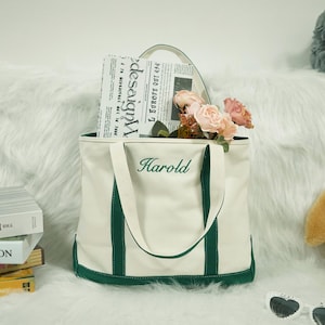 Peut inclure: Sac fourre-tout crème avec des détails verts et le nom "Harold" brodé sur le devant. Le sac contient un journal et des fleurs. Une pile de livres est à gauche. Le sac a une poignée blanche.