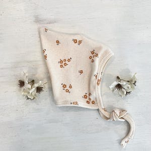 Pode incluir: Um gorro de bebê creme com uma textura canelada e um padrão de pequenos desenhos florais marrons. O gorro tem um fecho de laço e é exibido em uma superfície de madeira branca. Dois pequenos detalhes florais estão em cada lado.