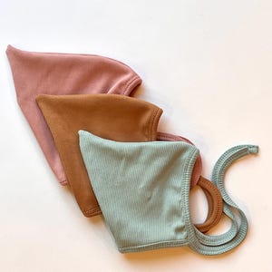 May include: Three ribbed bonnets in dusty rose, terracotta, and sage green. Each bonnet features a pointed top, a rounded face opening, and long fabric ties attached to the sides for securing the head covering in place.
