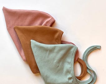 May include: Three ribbed bonnets in dusty rose, terracotta, and sage green. Each bonnet features a pointed top, a rounded face opening, and long fabric ties attached to the sides for securing the head covering in place.