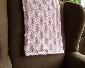 Op de afbeelding: Een roze gehaakte babydeken met een ruitpatroon en een textuur bobbelsteek. De deken is gedrapeerd over de rugleuning van een bruine fauteuil.