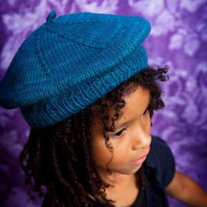 May include: A young person with long dark hair wearing a teal blue knitted beret and a dark blue shirt.