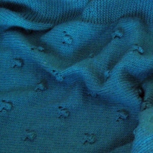 May include: A close-up of a teal blue knitted fabric with a textured pattern. The fabric is soft and has a slightly fuzzy texture.