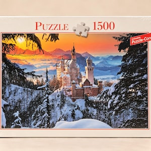 Peut inclure: Une boîte de puzzle de 1500 pièces représentant un château dans des montagnes enneigées au coucher du soleil. La boîte porte l'inscription Puzzle 1500 et une bannière rouge avec le texte inclusive Puzzle-Conserver dans le coin supérieur droit.