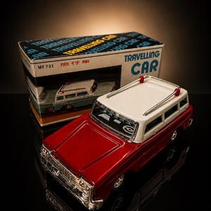 May include: A red and white toy car with a matching box. The car has a white roof and silver detailing. The box reads "TRAVELLING CAR" in blue and yellow text. The car is a vintage model.