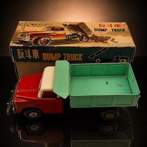 Könnte beinhalten: Vintage Spielzeug-Muldenkipper mit roter Kabine, weißem Dach und grünem Bett. Der LKW ist aus Metall und hat schwarze Räder. Die Originalverpackung befindet sich hinter dem LKW und zeigt eine Zeichnung des LKW. Auf der Box stehen die Worte "DUMP TRUCK" auf Englisch und Chinesisch.