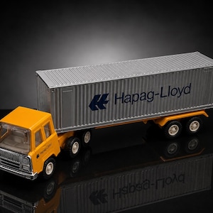 Könnte beinhalten: Ein gelber und silberner Spielzeug-LKW mit einem Container-Anhänger. Der Container ist silberfarben und trägt den blauen Schriftzug "Hapag-Lloyd". Der LKW hat eine gelbe Kabine und einen silbernen Kühlergrill. Die Räder sind schwarz mit weißen Felgen.