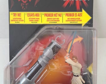May include: A Star Wars Episode I Deluxe Obi-Wan Kenobi action figure in original packaging. The card features Darth Maul, the text Try Me, and a plastic bubble containing a small Obi-Wan figure with a blue lightsaber and a large silver lightsaber hilt.