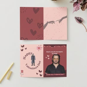 May include: A pink and red Valentine's Day card with a man in a black shirt and glasses. The text on the card reads "Won't you fix my heart strings & be my Valentine?"