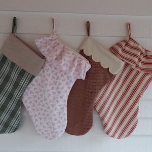 May include: Four fabric Christmas stockings hanging in a row. The first is green plaid with a tan cuff. The second is white with a small pink bow pattern and a ruffled top. The third is brown gingham with a cream scalloped cuff. The fourth is red and white striped with a ruffled top.