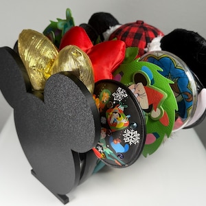 May include: A black Mickey Mouse-shaped stand with multiple pairs of colorful Disney character ear headbands. The headbands feature various characters, including Mickey Mouse, Minnie Mouse, and others.
