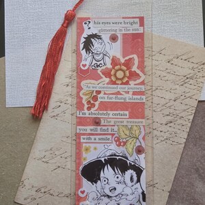 May include: A handmade bookmark with a red tassel. The bookmark features anime-style illustrations and text, including the phrases "his eyes were bright" and "you will find it with a smile." Decorative floral accents and a clear protective layer are also present.