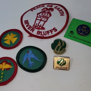 K&ouml;nnte beinhalten: Eine Sammlung von Pfadfinderinnen-Abzeichen und Anstecknadeln. Die Aufn&auml;her umfassen einen wei&szlig;en und roten Aufn&auml;her mit dem Text "PEACE ON EARTH" und "RIVER BLUFFS" sowie mehrere runde Aufn&auml;her mit verschiedenen Designs. Es gibt auch eine gr&uuml;ne Anstecknadel.