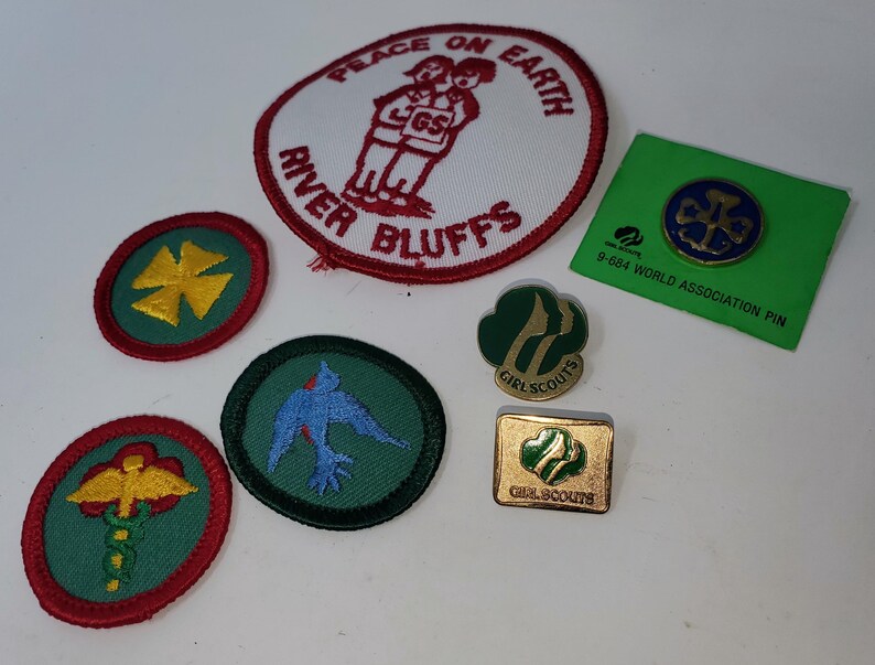 K&ouml;nnte beinhalten: Eine Sammlung von Pfadfinderinnen-Abzeichen und Anstecknadeln. Die Aufn&auml;her umfassen einen wei&szlig;en und roten Aufn&auml;her mit dem Text "PEACE ON EARTH" und "RIVER BLUFFS" sowie mehrere runde Aufn&auml;her mit verschiedenen Designs. Es gibt auch eine gr&uuml;ne Anstecknadel.