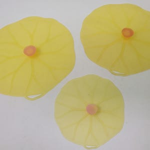 May include: Three yellow silicone bowl covers shaped like lily pads, each with a small pink center. The covers vary in size and have a leaf-like design. A small loop is visible on each cover.
