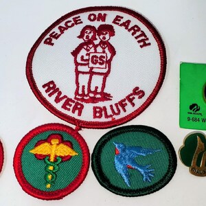 K&ouml;nnte beinhalten: Eine Sammlung von Pfadfinderinnen-Abzeichen und Anstecknadeln. Die Abzeichen umfassen ein gro&szlig;es rundes Abzeichen mit dem Text "PEACE ON EARTH" und "RIVER BLUFFS" sowie mehrere kleinere Abzeichen mit verschiedenen Designs. Es gibt auch mehrere Anstecknadeln, darunter eine gr&uuml;ne Anstecknadel mit einem goldenen Emblem.