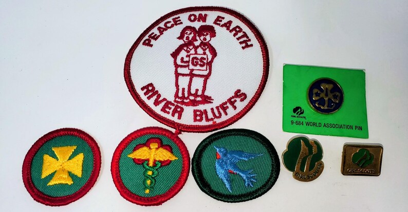 K&ouml;nnte beinhalten: Eine Sammlung von Pfadfinderinnen-Abzeichen und Anstecknadeln. Die Abzeichen umfassen ein gro&szlig;es rundes Abzeichen mit dem Text "PEACE ON EARTH" und "RIVER BLUFFS" sowie mehrere kleinere Abzeichen mit verschiedenen Designs. Es gibt auch mehrere Anstecknadeln, darunter eine gr&uuml;ne Anstecknadel mit einem goldenen Emblem.