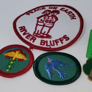 K&ouml;nnte beinhalten: Eine Sammlung von Girl Scout-Aufn&auml;hern und -Anstecknadeln. Die Aufn&auml;her umfassen einen gro&szlig;en ovalen Aufn&auml;her mit dem Text "Peace on Earth" und "River Bluffs" sowie mehrere kleinere Aufn&auml;her mit verschiedenen Designs. Es gibt auch mehrere Anstecknadeln, darunter eine World Association-Anstecknadel.