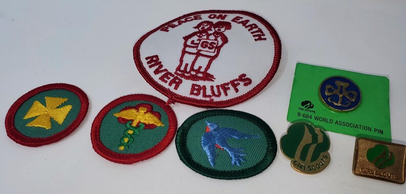 K&ouml;nnte beinhalten: Eine Sammlung von Girl Scout-Aufn&auml;hern und -Anstecknadeln. Die Aufn&auml;her umfassen einen gro&szlig;en ovalen Aufn&auml;her mit dem Text "Peace on Earth" und "River Bluffs" sowie mehrere kleinere Aufn&auml;her mit verschiedenen Designs. Es gibt auch mehrere Anstecknadeln, darunter eine World Association-Anstecknadel.